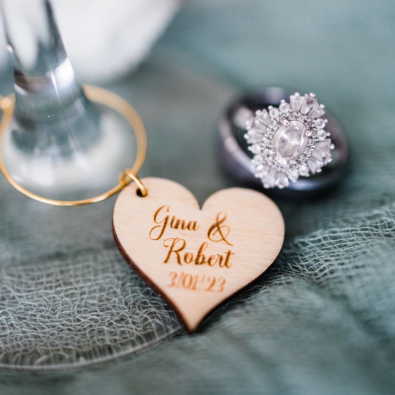 May include: A wooden heart-shaped wedding favor with the names Gina & Robert and the date 3/01/23 engraved on it. A silver ring with a large diamond is in the background.