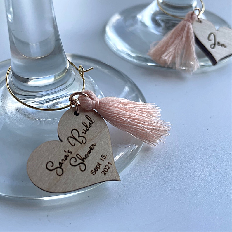 May include: A wooden heart-shaped tag with the text "Sara's Bridal Shower Sept 15, 2021" and a pink tassel attached to a gold wire hoop.
