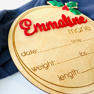 May include: A round wooden plaque with a holly sprig and berries in the top left corner. The name "Emmaline marie" is written in red letters. The plaque has spaces to fill in the date, time, weight in pounds, and length.