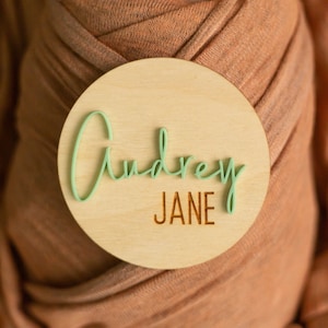 May include: A round wooden sign with the name "Audrey Jane" written in green cursive lettering on a light wood background.