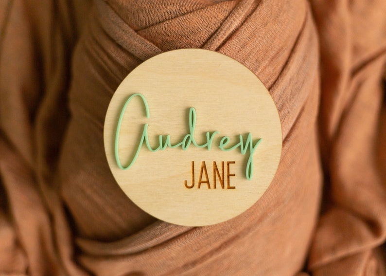 May include: A round wooden sign with the name "Audrey Jane" written in green cursive lettering on a light wood background.