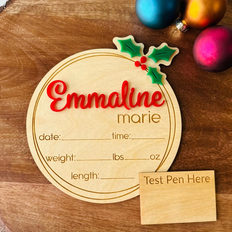 May include: A round wooden plaque with a gold border and a holly sprig design. The plaque is decorated with the name "Emmaline marie" in red lettering. It has spaces to fill in the date, time, weight in pounds and ounces, and length.