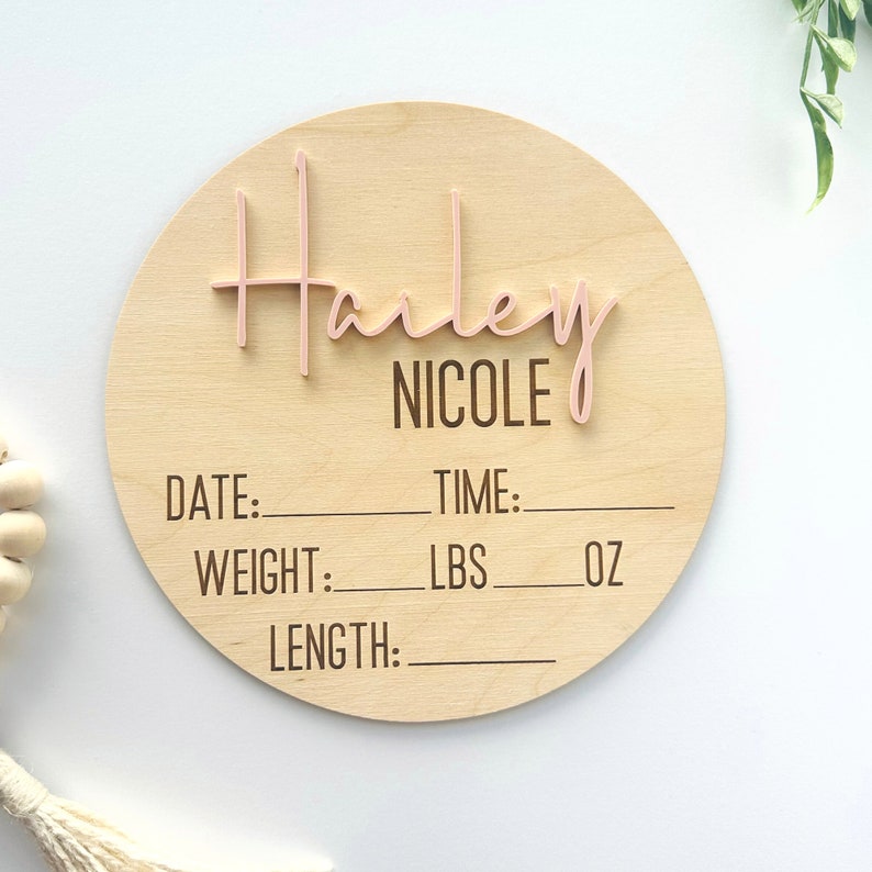 May include: A round wooden baby announcement plaque with the name "Harley Nicole" in pink script. The plaque has spaces to fill in the baby's date, time, weight in pounds and ounces, and length.