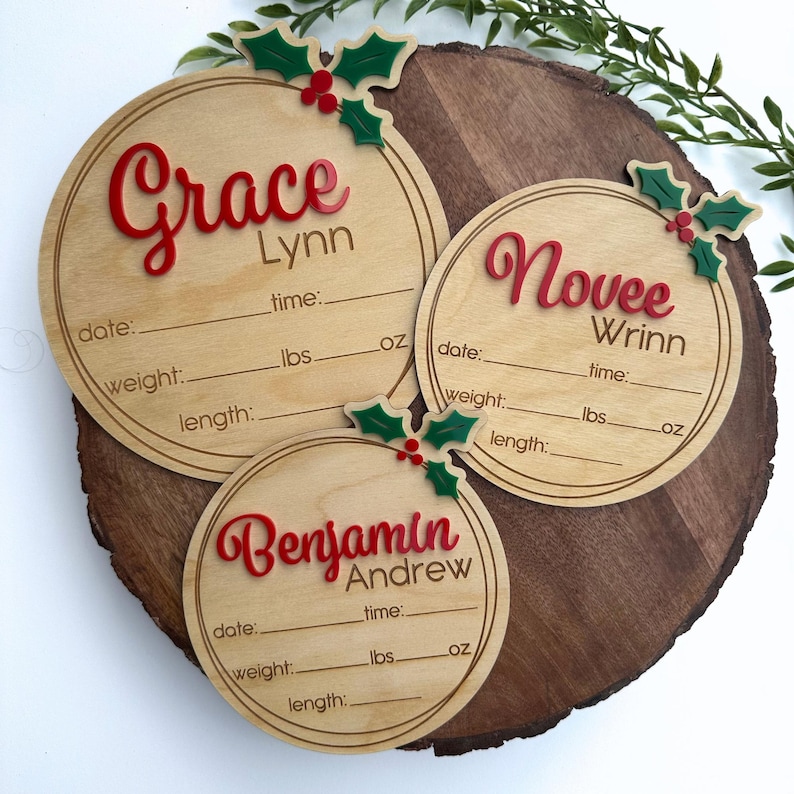 May include: Three round wooden ornaments with holly and berry accents. Each ornament has a name, "Grace Lynn", "Novee Wrinn", and "Benjamin Andrew", with spaces for date, time, weight in pounds and ounces, and length.