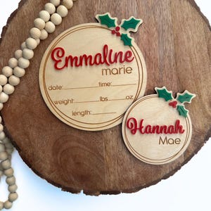 May include: Two wooden Christmas ornament-style birth announcement plaques with holly and berry accents. The top plaque reads "Emmaline marie" with spaces for date, time, weight in pounds and ounces, and length. The bottom plaque reads "Hannah Mae".