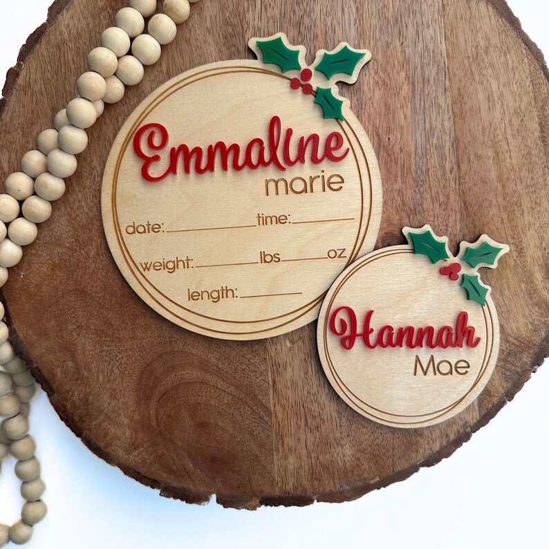 May include: Two wooden Christmas ornament-style birth announcement plaques with holly and berry accents. The top plaque reads "Emmaline marie" with spaces for date, time, weight in pounds and ounces, and length. The bottom plaque reads "Hannah Mae".