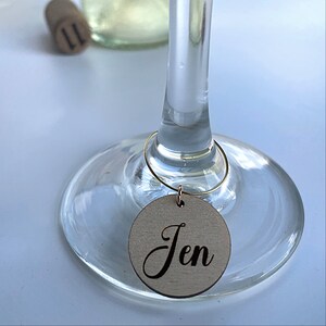 May include: A wooden wine glass charm with the name "Jen" engraved on it. The charm is attached to a gold wire ring.