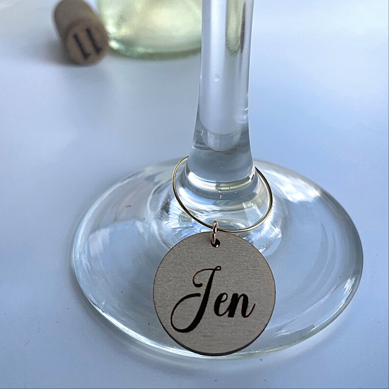 May include: A wooden wine glass charm with the name "Jen" engraved on it. The charm is attached to a gold wire ring.