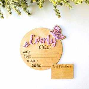 May include: A wooden baby announcement plaque with the name "Everly Grace" and two pink butterflies. The plaque has spaces to write the date, time, weight, and length. A separate wooden plaque says "Test Pen Here".