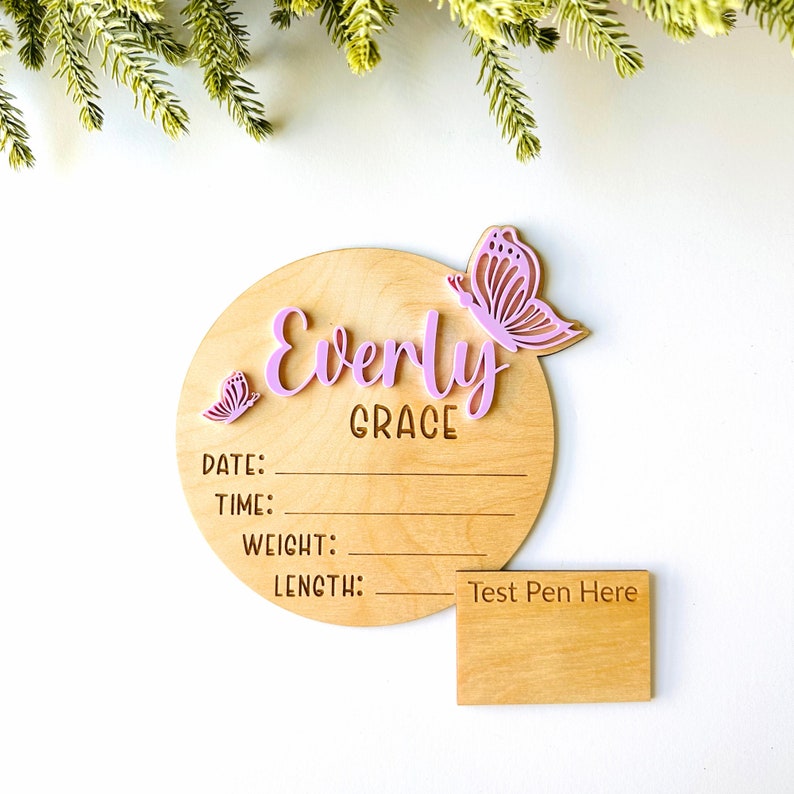May include: A wooden baby announcement plaque with the name "Everly Grace" and two pink butterflies. The plaque has spaces to write the date, time, weight, and length. A separate wooden plaque says "Test Pen Here".