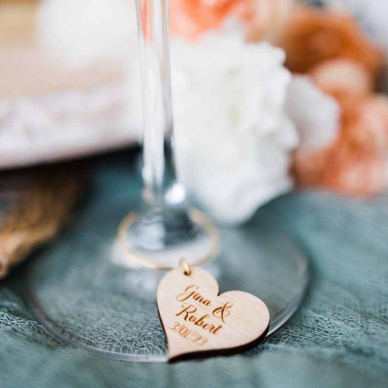 May include: A wooden heart-shaped tag with the text "Gina & Robert 3.01.23" engraved on it. The tag is attached to a glass wine glass.