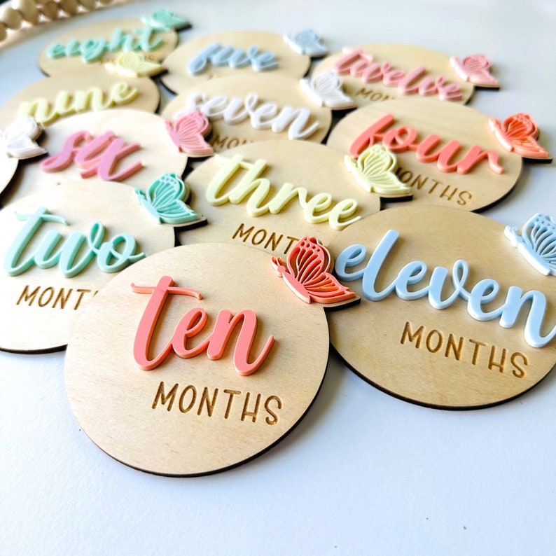 May include: A set of 12 wooden baby milestone discs with pastel coloured butterflies and numbers from one to twelve. Each disc has the word "months" engraved on it.