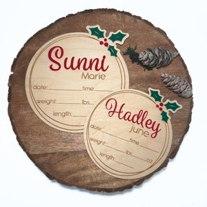 May include: Two round wooden plaques with holly and berry decorations. The top plaque reads "Sunni Marie" with spaces for date, weight, length, and time. The bottom plaque reads "Hadley June" with spaces for date, weight, length, and time.