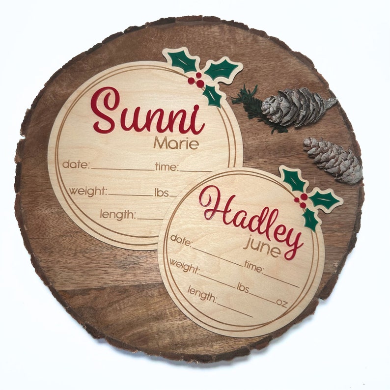 May include: Two round wooden plaques with holly and berry decorations. The top plaque reads "Sunni Marie" with spaces for date, weight, length, and time. The bottom plaque reads "Hadley June" with spaces for date, weight, length, and time.
