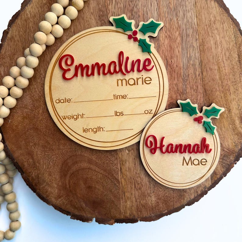 May include: Two round wooden ornaments with holly and berry accents. The larger ornament has the text "Emmaline marie" and spaces for birth information, including date, time, weight in pounds and ounces, and length. The smaller ornament has the text "Hannah Mae".
