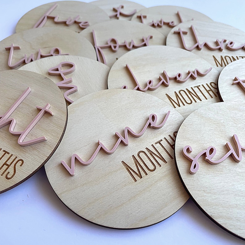 May include: A set of 11 wooden baby milestone discs with pink script numbers and the word "months" engraved on each disc. The discs are arranged in a pile, with the number "nine" visible in the center.