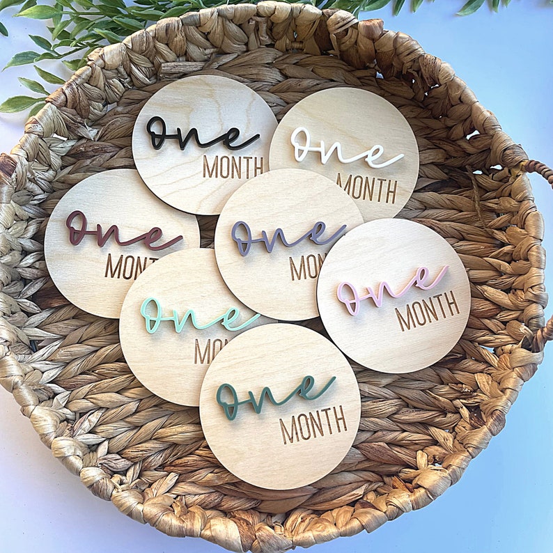 May include: Set of six wooden baby milestone discs with the word "one" and "month" written on each disc. The discs are in a wicker basket and feature different colored acrylic lettering.
