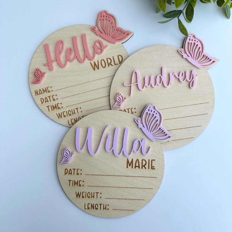 May include: Three wooden baby announcement plaques with pink butterfly accents. The plaques have spaces to write the baby's name, date, time, weight, and length. The plaques read "Hello World", "Audrey", and "Willa Marie".