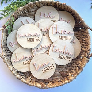 May include: A set of 12 wooden baby milestone discs with pink script numbers and the word "MONTHS" on each disc. The discs are arranged in a wicker basket.
