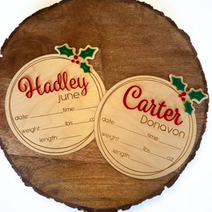 May include: Two round wooden ornaments with holly accents and spaces to fill in baby's name, date, time, weight, and length. The ornaments are decorated with red and green holly and the names "Hadley June" and "Carter Donavon" are written in red.