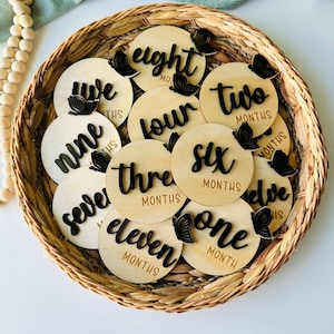 May include: A set of twelve wooden baby milestone discs with black butterfly accents and the numbers one through twelve written on them. Each disc has the word "months" written below the number.