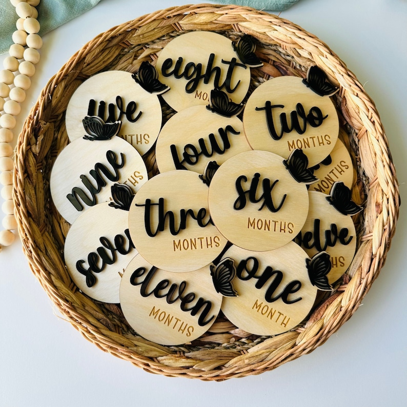 May include: A set of twelve wooden baby milestone discs with black butterfly accents and the numbers one through twelve written on them. Each disc has the word "months" written below the number.