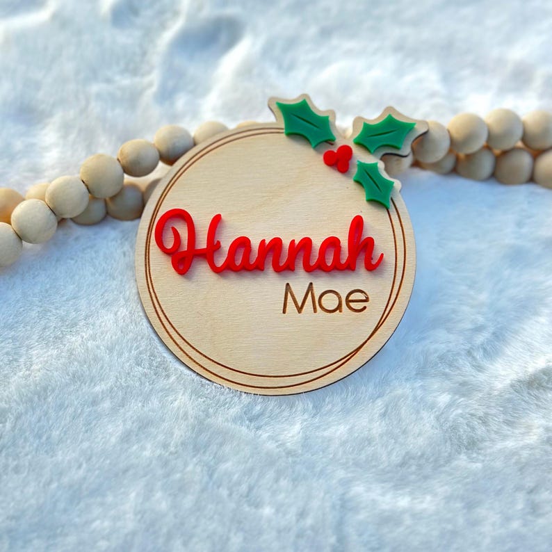 May include: A round wooden ornament with a red "Hannah Mae" name and a green holly sprig with red berries.