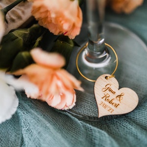 May include: A wooden heart-shaped wedding favor tag with the text "Gina & Robert 3/01/23" engraved on it. The tag is attached to a gold wire ring.