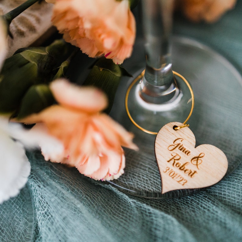 May include: A wooden heart-shaped wedding favor tag with the text "Gina & Robert 3/01/23" engraved on it. The tag is attached to a gold wire ring.