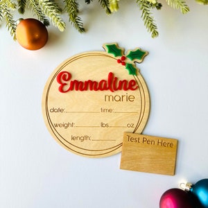 May include: A round wooden ornament with a holly design and the name "Emmaline marie" written in red. The ornament has spaces to record the date, time, weight in pounds and ounces, and length in inches. A separate wooden rectangle with the text "Test Pen Here" is included.