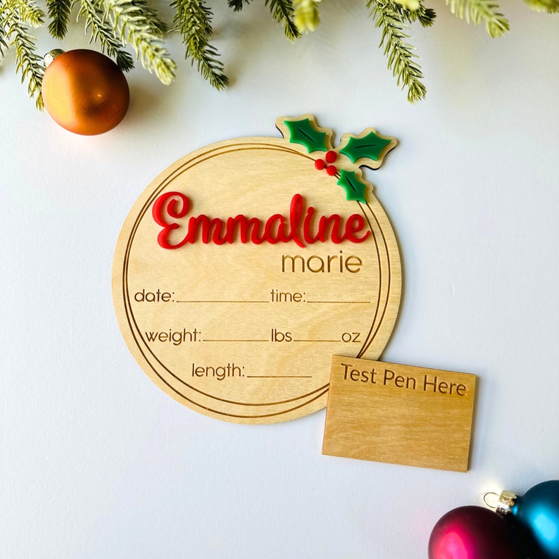 May include: A round wooden ornament with a holly design and the name "Emmaline marie" written in red. The ornament has spaces to record the date, time, weight in pounds and ounces, and length in inches. A separate wooden rectangle with the text "Test Pen Here" is included.