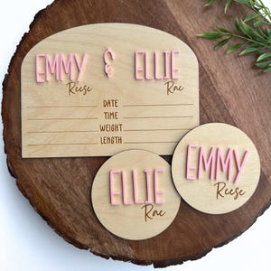 May include: Two wooden baby name plaques with pink lettering. One plaque has the names "Emmy Reese" and "Ellie Rae" and a space to record birth information. The other plaque is round and says "Emmy Reese".