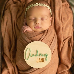 May include: A newborn baby girl is sleeping in a brown swaddle blanket. A wooden circle with the name "Audrey Jane" written in green and brown is placed on the blanket.
