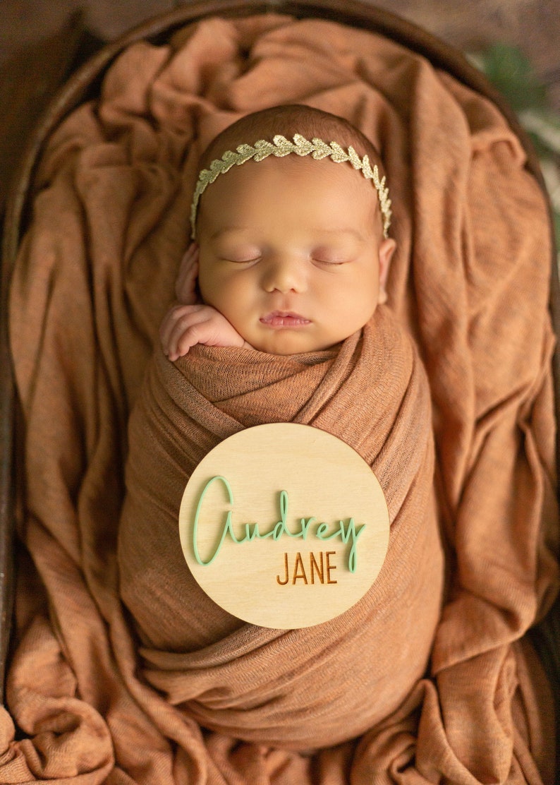 May include: A newborn baby girl is sleeping in a brown swaddle blanket. A wooden circle with the name "Audrey Jane" written in green and brown is placed on the blanket.