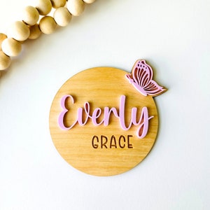 May include: A round wooden sign with the name "Everly Grace" written in pink letters and a pink butterfly cutout on the top right corner.