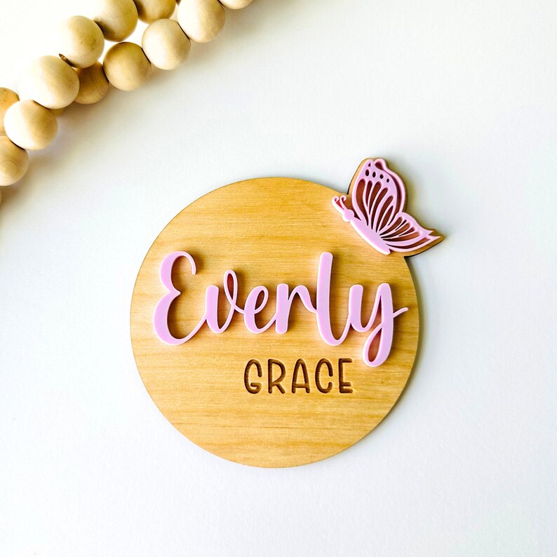 May include: A round wooden sign with the name "Everly Grace" written in pink letters and a pink butterfly cutout on the top right corner.