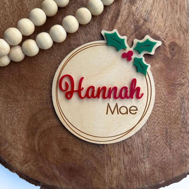 May include: A round wooden ornament with a red and green holly design and the name "Hannah Mae" written in red cursive.