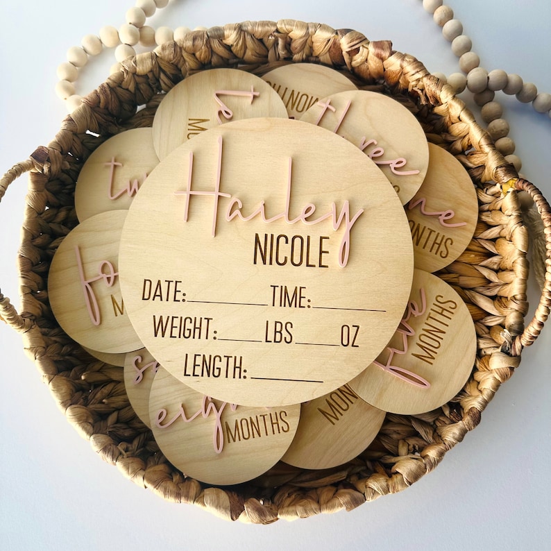 May include: A set of wooden baby milestone discs with pink lettering. The largest disc has the name "Harley Nicole" and spaces to record the baby's date, time, weight in pounds and ounces, and length. The other discs show the numbers one through twelve, with the word "months" written on each.