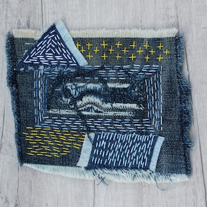 May include: A blue denim fabric patch with various embroidered designs in yellow and blue thread. The patch is frayed around the edges and has a distressed look.