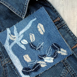 May include: A blue denim jacket with a patch of blue denim sewn on the front. The patch has a design of a tree with white branches and blue and white leaves.