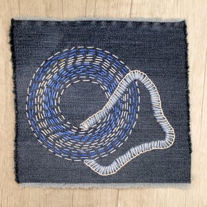 Könnte beinhalten: Quadratischer Denim-Patch mit ausgefranstem Rand. Der Patch zeigt ein gesticktes Design in Blau- und Weißtönen, das eine strukturierte, abstrakte Form erzeugt. Der Hintergrund ist ein mittelblau gewaschener Denim.