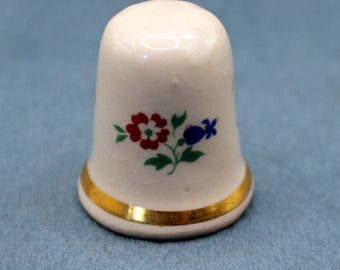 May include: White ceramic thimble with a gold trim and a floral design featuring red, white, blue, and green flowers.
