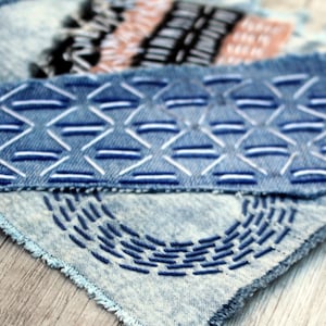Könnte beinhalten: Hellblaue, säuregewaschene Jeansstoffreste mit handgestickten Mustern. Ein Stück zeigt ein geometrisches Design aus weißem und blauem Garn, ein anderes ein bogenförmiges Muster in Dunkelblau und ein drittes weist schwarze und hellbraune Stickereien auf.