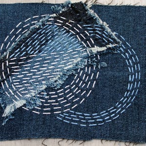 Puede incluir: Tela vaquera azul oscuro con costuras blancas y azul claro. El diseño presenta un parche rasgado y deshilachado con un patrón circular y líneas paralelas. La costura crea una apariencia texturizada y artesanal.