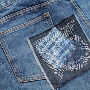 May include: Close-up of blue denim jeans with a decorative patch on the back pocket. The patch features white stitching in a geometric design and a frayed edge. The jeans have a classic five-pocket design.