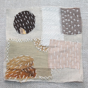 Könnte beinhalten: Ein beigefarbener Stoff-Patchwork mit verschiedenen Stichmustern. Der Stoff ist zu einer quadratischen Form zusammengenäht. Der Patchwork enthält einen braunen Kreis mit weißen Nähten, einen weißen Stoff mit weißen Nähten, einen beigefarbenen Stoff mit weißen Nähten und einen braunen Stoff mit weißen Nähten.