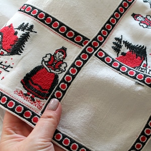 May include: White fabric with red and black embroidered designs. The designs include a woman in traditional dress, a house, and trees. The fabric has a black and red border with a repeating circle pattern.