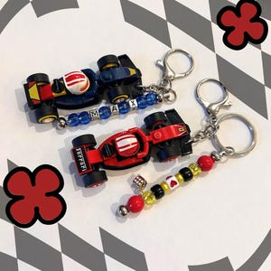 Puede incluir: Dos llaveros de coches de carreras de juguete. Uno es rojo y negro con una franja blanca, el otro es azul y amarillo. Ambos tienen llaveros plateados y detalles de cuentas. El coche azul tiene el nombre "MAX" escrito con cuentas.