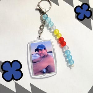 Könnte beinhalten: Ein klarer Acryl-Schlüsselanhänger mit einem Foto einer Person mit blauer Kappe. Der Schlüsselanhänger hat einen silbernen Verschluss und eine Perlenkette mit blauen, weißen, gelben und roten Perlen. Zwei blaue blütenförmige Aufkleber befinden sich im Hintergrund.