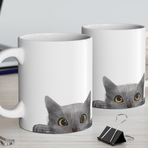 May include: Two white ceramic mugs with a gray cat peeking over the rim of each mug. The cat has yellow eyes and a gray nose.